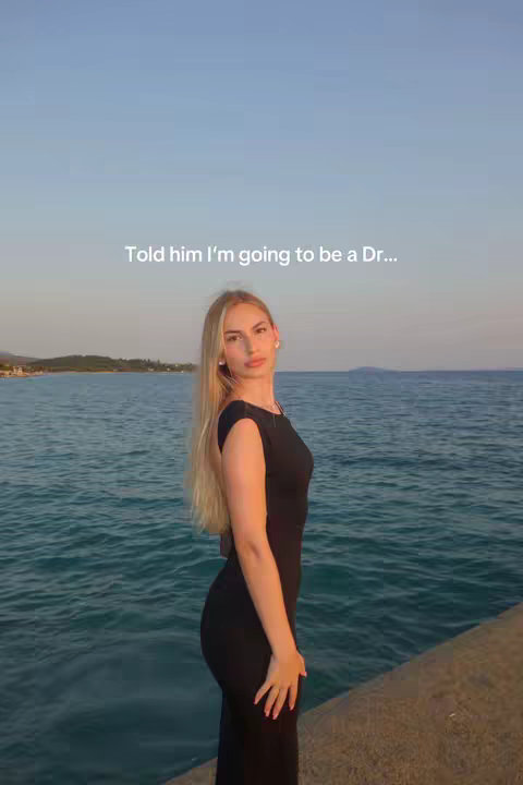A woman stands on a stone structure facing the camera, with a background of the ocean and a shoreline. The sky is a gradient of colors from a bright horizon to a lighter blue, indicating either sunrise or sunset. The woman is wearing a black dress, with long blonde hair and a relaxed pose. The image has a natural lighting, suggesting an outdoor setting.