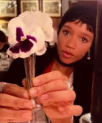 A woman holds a flower in a clear vase, angled slightly towards the camera. She is wearing a black beanie, a black choker, and has earrings. Her face is the primary focus, with a soft, slightly out-of-focus background including framed pictures.