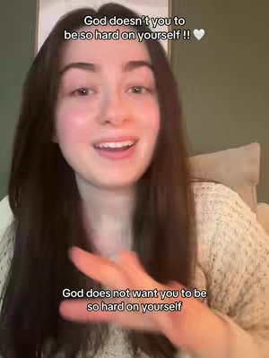 A woman with long dark hair is centered in the frame, smiling with her mouth open. Above her, text reads, 'God doesn't you to be so hard on yourself !! ", with a heart emoji. Below, the text states, 'God does not want you to be so hard on yourself.' The background is a soft, out-of-focus setting with a hint of green.