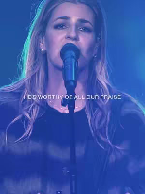 A woman with long blonde hair is singing into a microphone, with a soft, cool-toned blue light illuminating her face and the microphone. Text overlayed on the bottom of the image reads, 'HE'S WORTHY OF ALL OUR PRAISE'.