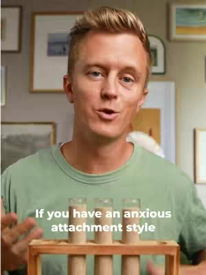 A man with blonde hair is speaking directly to the camera. He's wearing a green t-shirt against a backdrop with framed pictures. In the foreground, there's a small wooden stand with three clear tubes filled with a beige liquid.