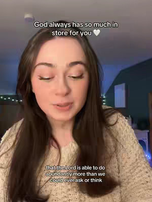 A young woman with long, dark hair is centered in the frame, speaking directly to the camera. She is wearing a cream-colored, textured sweater. Above her, a text overlay reads, "God always has so much in store for you," along with a white heart emoji. Behind her, the background is slightly blurred, featuring glimpses of a dark wall and possibly a piece of furniture. The lighting appears to be soft and diffused.