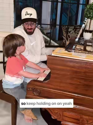 A middle-aged man wearing glasses and a hat with the word "Dang" on it is sitting at a piano, coaching a young child on how to play. The child is seated next to him, with the child's hands on the piano keys.