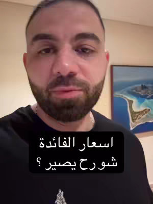 A selfie-style video image featuring a man looking directly at the camera. He has a beard and short hair. Overlaying his face is a black box with text in Arabic, posing a question about interest rates. Behind him is a light-colored wall and a framed picture with a blue landscape.