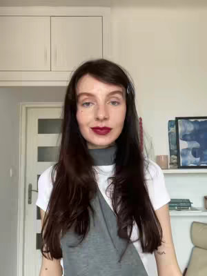 A woman with dark hair and burgundy lipstick is centered, facing the camera. She is wearing a white t-shirt and a gray sleeveless top. The background suggests a home setting with a cabinet and artwork.