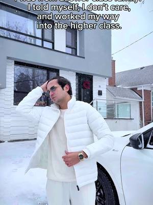 A man in a white puffer jacket and sunglasses poses in front of a modern house with snow on the ground. A white car is parked next to him.