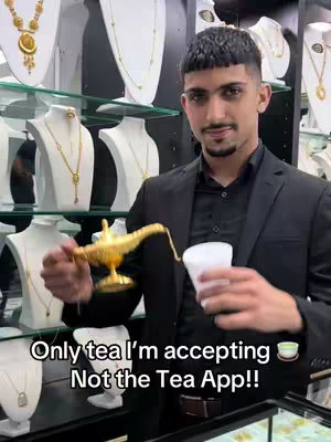 A man in a black suit stands in a jewelry store, holding a golden lamp and a white cup. The background displays a variety of necklaces on white mannequins. Text overlay at the bottom suggests rejecting a "Tea App" in favor of real tea.