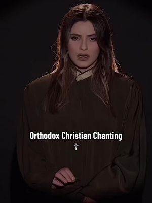 A medium shot of a young woman with long brown hair and a neutral expression, wearing a dark brown robe-like garment with a lighter colored accent at the neckline, against a dark background. Text overlays are 'Orthodox Christian Chanting' and a small cross icon. The lighting appears to be focused on the woman.