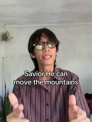 A man with dark hair and glasses is centered in the frame with a blurred background. He is wearing a striped shirt and pointing both index fingers out towards the viewer. Text overlayed on the image says 'Savior He can move the mountains'.