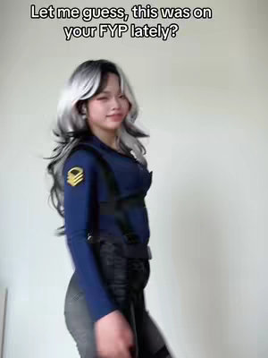 A young woman with gray and black hair stands in front of a white background, angled slightly to the side. She wears a dark blue uniform with a high collar and straps. The overlay text reads, "Let me guess, this was on your FYP lately?"