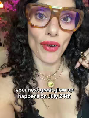 A woman with curly dark hair, wearing large square glasses, and red lipstick is looking directly at the camera. She has layered necklaces, and is in a lit room. Text at the bottom of the image says 'your next great glow up happens on July 24th'.