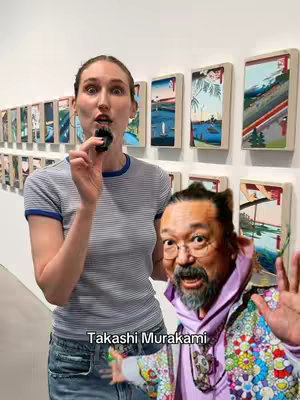 A woman stands in front of a wall gallery display. A smaller image cropped in presents a photo of artist Takashi Murakami in close proximity. The woman facing the camera holds a small black item up to her mouth. The background presents various pieces of what appears to be artwork.