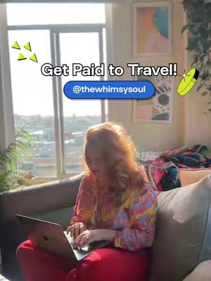 A woman sits on a couch near a window, working on a laptop. Text overlays read: 'Get Paid to Travel!' and '@thewhimsysoul.' The scene is brightly lit, suggesting a sunny day, with the woman and text primarily displayed against background of windows and indoor space.