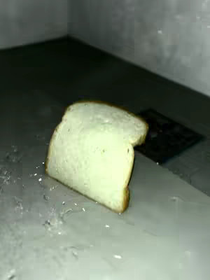 A slice of white bread is positioned slightly off-center on a light-colored surface, with a dark, blurred area in the background. The bread has a golden-brown crust on its edges. There are tiny water droplets scattered on the surface near the bread.