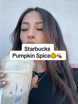 A woman with long dark hair and closed eyes is holding a clear plastic cup filled with a light brown beverage. The drink is likely a latte or coffee, and it has a creamy appearance. The woman appears to be in the middle of drinking the beverage, and she's positioned against an out-of-focus background, possibly outdoors.