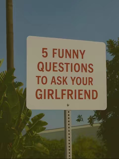 A photograph shows a white rectangular sign with rounded corners on a metal pole. The sign has large, red text that reads, "5 FUNNY QUESTIONS TO ASK YOUR GIRLFRIEND". The background includes a pale blue sky, leafy green trees, and glimpses of buildings.