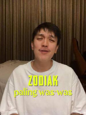 A man with short dark hair and a mustache is centered in the frame, wearing a white t-shirt. The background is a blurred brown vertical striped pattern. There is golden yellow text overlaying the image that says, "ZODIAK paling was-was".