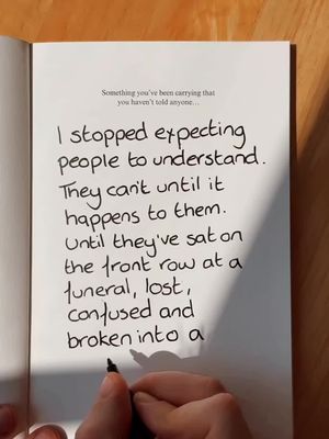 The image displays an open book on a wooden surface, with handwritten text and a hand holding a pen. The text fills the majority of the right page and the background is a warm tone.