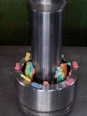 The image shows a large, cylindrical metal device, possibly an industrial press or machine, centered on a surface. The device has a wide, circular base with a central opening. Inside the base, colorful gummy candies are arranged around the central cylinder. As the image is captured it shows the pressure of the metal object on the gummy bears. The overall composition is focused and visually appealing.