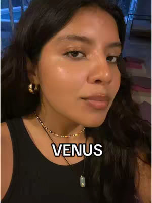 A close-up captures a woman with dark hair and a natural makeup look, set against a blurred background. She wears gold hoop earrings and a necklace. Overlaid on the image is the word 'VENUS'.
