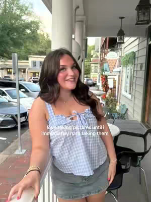 A young woman with dark hair smiles while standing on a porch. She wears a blue and white gingham top and a denim skirt. The background shows a street with cars, buildings, and a wooden porch with chairs and tables.