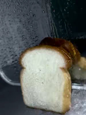 A slice of white bread is positioned in front of a partly obscured, slightly out-of-focus background. Water droplets are visible running down the glass on the upper part of the frame. The bread itself has a lightly toasted brown crust around the edges and is glistening, as if wet.