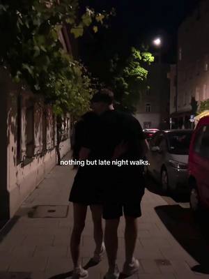 Two figures are embracing on a dimly lit sidewalk at night. Streetlights and tree branches are overhead, with buildings and parked cars in the background. Text overlayed.