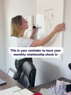 A woman is standing in front of a white wall, holding a thin black marker. She appears to be interacting with a framed piece of art depicting a line drawing of a person reading a book. The artwork is mounted against the wall. The composition suggests a home interior, possibly an office or living room, with natural lighting. A text overlay sits in the lower part of the image and reads "This is your reminder to have your monthly relationship check in."