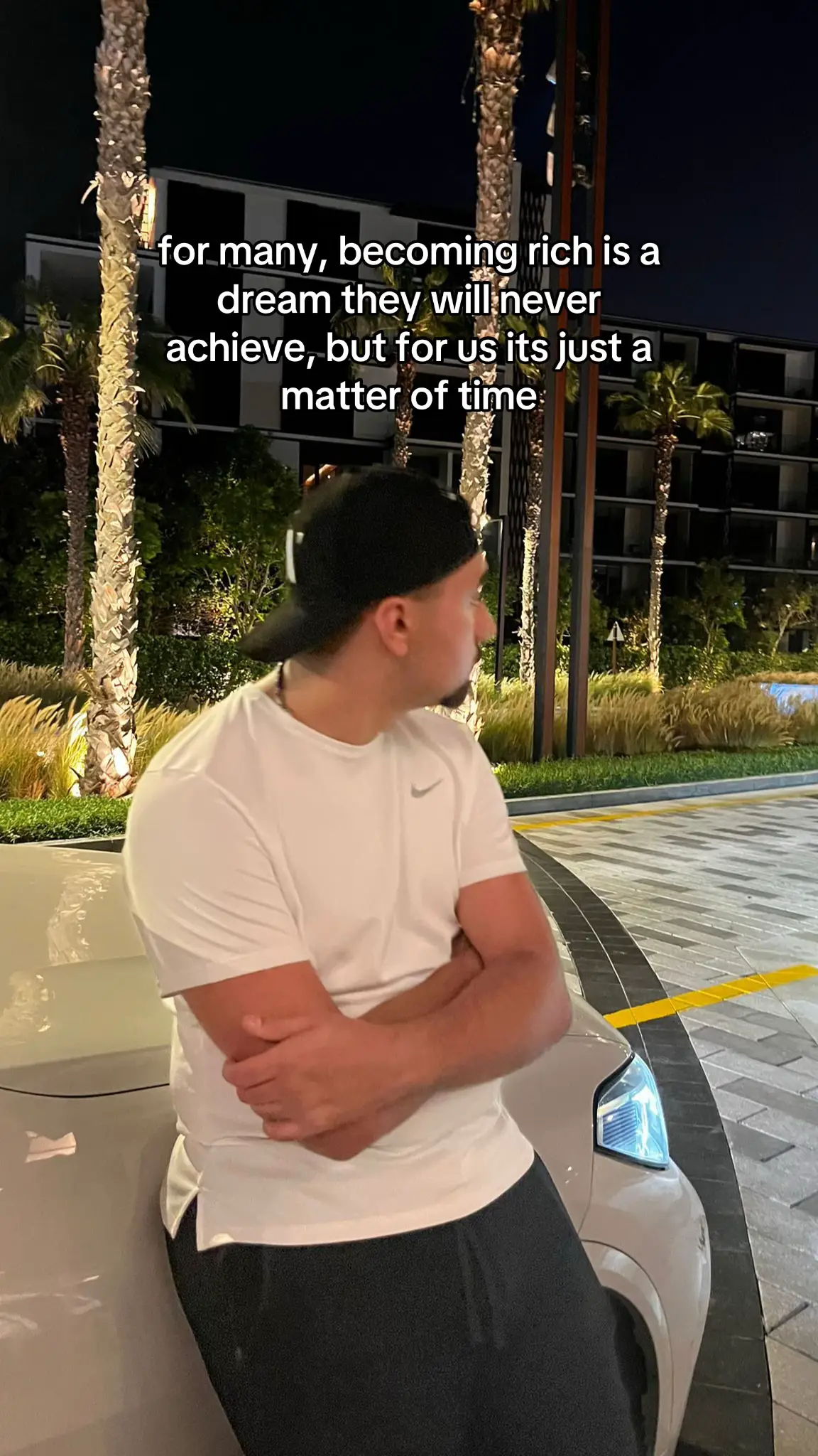 A man in a white t-shirt and black cap is leaning against a car at night. Text overlayed reads: "for many, becoming rich is a dream they will never achieve, but for us its just a matter of time."
