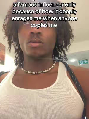 A selfie-style shot of a young Black man from the chest up. He is wearing a white tank top and a beaded necklace. His hair is dark and curly. The background is blurred, but appears to be a hallway or interior space. There is text overlaid at the top of the image.