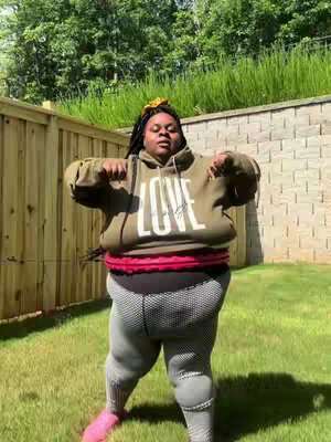 A full-body shot of a person standing outside on a sunny day, wearing a green hoodie with the word "LOVE" across the front, dark leggings with a mesh design, and pink shoes. The person is standing in front of a wooden fence on a grassy lawn, with a concrete wall behind them. They are wearing a floral headband and have their arms up, as if working out.