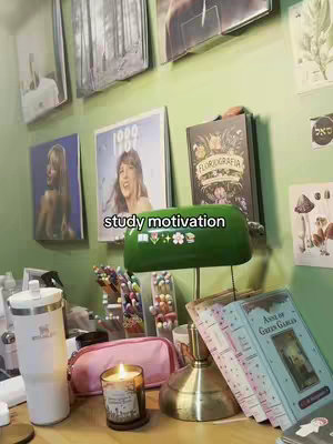 A well-lit indoor scene showcasing a desk setup, with several books including "Anne of Green Gables" and "Florigrafia", a lit candle, a water bottle, a pink pencil case, and other stationery items. Artwork lines the wall in the background, and a green lamp provides additional light.