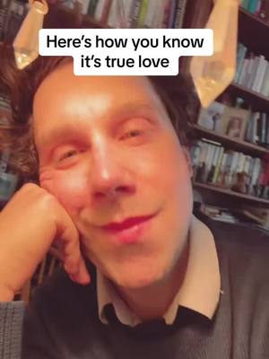 A man with auburn hair is looking at the camera and smiling. The background features a bookshelf.