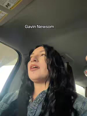 A selfie from inside a car. The woman in the image is speaking to the camera. Text that reads 'Gavin Newsom' appears over her head. The interior of the car is visible, as is part of the visor and windshield.