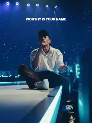 A man in a white shirt and black baseball cap sits cross-legged on a ledge with a dark background, raising one hand to his face as if holding a microphone. Text at the top reads, 'Worthy is your name'.