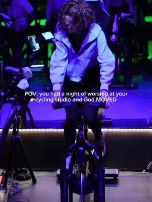 A person sits on a stationary bike in a spin studio with a large light in the background. The frame is in a dim setting with focused light and adds text overlays.