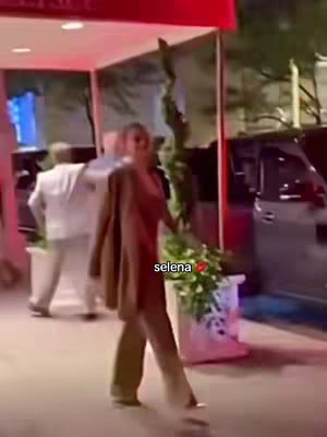 A woman in a brown outfit walks on a sidewalk and near a building entrance. The text "Selena" with a heart appears on the image.