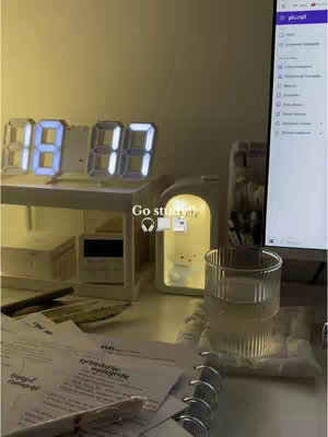 A well-lit desk setup featuring a digital clock displaying '18:00', a small illuminated bear lamp, a glass of water, and study materials. The background is a muted yellow, creating a warm, inviting ambiance. The composition is tidy and focuses on items related to studying.
