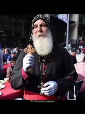 A man in religious garb, holding a cross, is shown in a crowd. The background is blurred, suggesting an outdoor setting. The man has a long white beard, a black hood, and a cross necklace. He is wearing the formal dress of his profession and white gloves. The images are taken from a medium-level distance that captures the surrounding, and at the bottom, there is the username added in the text