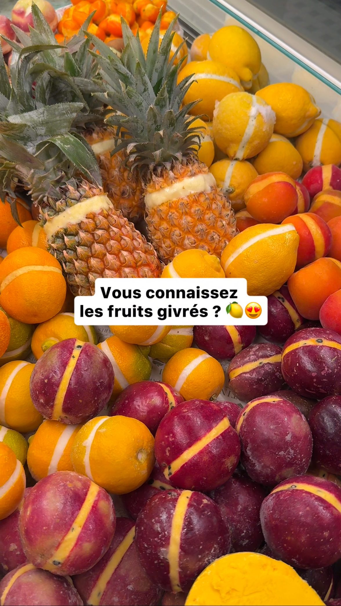 An overhead shot showcases a colorful display of various fruits. The foreground is filled with round, reddish-purple fruits, each marked with a yellow stripe. Interspersed are bright yellow lemons and oranges, also with painted stripes. Behind, the image features peeled pineapples along with another arrangement of lemons and oranges. The fruits are closely packed, creating a vibrant and appetizing visual.