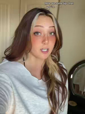 A woman with long, multi-toned hair is centered in the frame, looking directly at the camera. She has a slight smile and is wearing a white v-neck shirt. The background is blurred, suggesting an indoor setting, possibly a home, with a glimpse of a dark-colored object to the right, which may be a mirror of some sort, in addition to white wall panels.