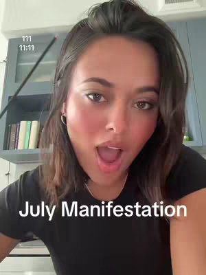 A selfie-style shot featuring a woman with dark hair and fair skin. She has expressive eyes and is positioned with her mouth open in an 'O' shape. The background is a domestic interior with neutral-colored cabinets, and some books are visible. Text on the image reads 'July Manifestation' in a clear, white font, and figures 111 and 11:11 are also visible on the upper left.