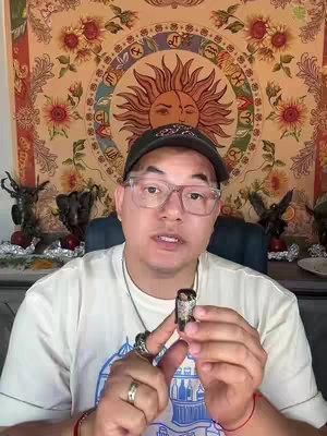A man with glasses is centered in the frame, holding a small, cylindrical dark object in his hands. He is wearing a white t-shirt and a black cap with a logo. Behind him, a colorful tapestry with a sun and zodiac signs hangs. The background gives a spiritual and artistic context to the video. The lighting is bright, and the overall composition is well balanced.