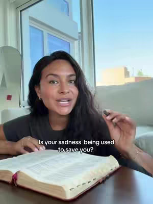 A woman with dark hair and fair skin is speaking directly to the camera while holding a small object. Behind her is a window with daylight and architectural details, as well as a neutral-toned couch. In front of her is an open book on a wooden surface.