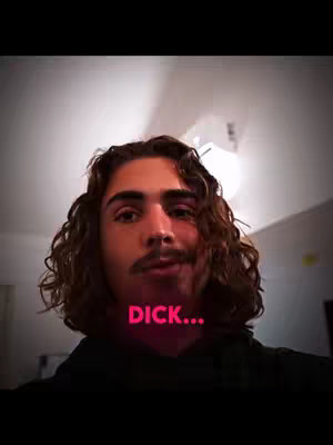 A man with long curly brown hair and a mustache is centered in the frame. The background is a lightly lit indoor space. Overlayed in the center is a pink rectangle with the word "DICK..." in capital letters.