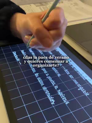 A hand with a teal-colored stylus is shown writing on a gray tablet screen which is situated on a desk. The screen displays text and a grid pattern. The background shows a white surface. The overall composition focuses on a close-up of writing activity.