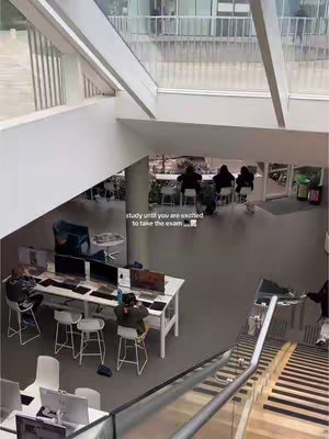 An overhead shot of a modern university building's interior. The image captures students in a brightly lit space with large windows. Students are seated at desks with computers, engaged in study. A set of stairs leads to a lower level where more students are at tables.