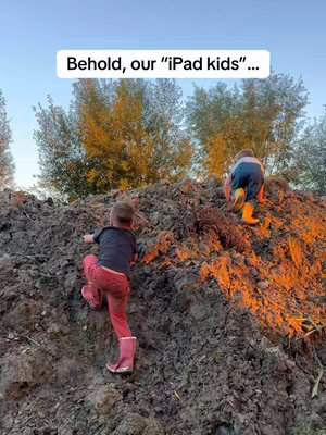 Two young children are climbing up a large pile of brown soil in a natural outdoor setting. The child in the foreground is seen from the back mid-climb, wearing pink boots and pink pants with a black t-shirt. The other child is further up the pile, also climbing. The sky is a pale blue, with trees in the background. The lighting is warm, suggesting either sunrise or sunset. A text overlay reading "Behold, our "iPad kids"...", is positioned centrally at the top of the image.