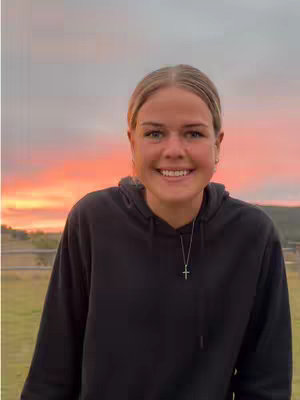 A woman wearing a black hoodie is smiling at the camera. The background shows a reddish-orange sunset and a hint of a field with a fence.