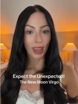 A woman with long, dark hair is centered in the frame, looking directly at the camera with her mouth slightly open and a warm expression. She's wearing a white top, and the background features warm-toned lighting, suggesting a bedroom setting with lamps visible.